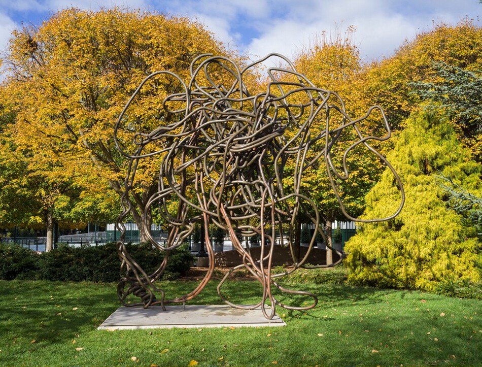 This sculpture features long looping forms resembling thick cords or vines that overlap with each other in a knotted jumble. It is made of a metallic material with a bronze-like finish, set on a flat rectangular base in an outdoor setting surrounded by green grass and trees with yellow and green foliage.