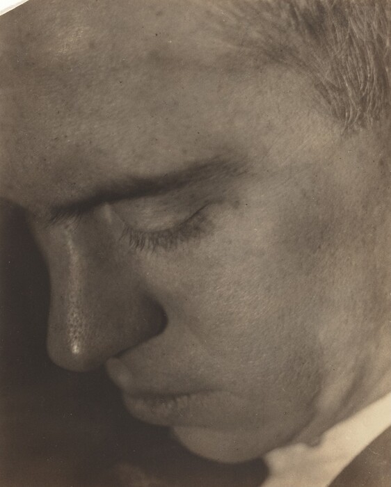 The image shows a close-up portrait of a person's face, focusing on the left side. The person's face is tilted downwards, showing a part of their forehead, closed eye, nose, and mouth. The facial features are in soft focus with a subtle texture on the skin and light eyelashes above the closed eyelid. The person has short, light-colored hair. No clothing is visible, and the background is out of focus.