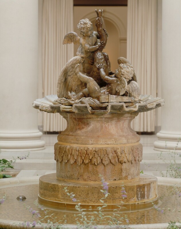 Two chubby, winged children hold onto a swan with its wings thrust back, beak open, and long neck curving up in this freestanding sculpture, which was originally a fountain cast in iron. The cherubs and swan are on a bunched-up cloth on a disk fluted like a seashell. In this photograph, the swan’s body is angled to our right. One cherub stands behind the swan to our left, and the other lies on his back with one hand and foot lifted, facing the swan. Both cherubs have caps of curly hair and short wings. Each has a strap across their chest, and a quiver with arrows lies next to the prone cherub. The sculpture rests on a pale peach-colored stone pedestal carved with a band of layered leaves around the middle. The base stands in a shallow basin of water. The fountain is photographed in a bright space with white marble columns and floors.