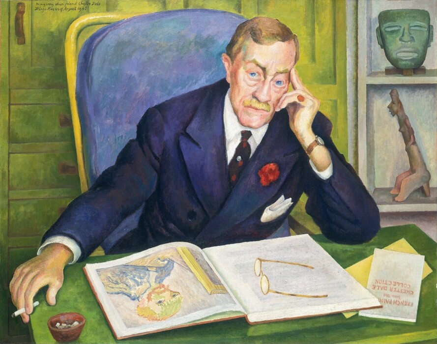 A pale-skinned man leans forward so his arms rest on the desk in front of us as he looks out with piercing blue eyes in this horizontal portrait painting. He rests his head in his left hand, to our right, with the first two fingers extended along his temple. His blond hair is carefully combed, and he has arched eyebrows, bags under his eyes, a wide nose, and a bristle mustache over his square, cleft chin. He wears a navy-blue suit jacket over a white button-down and dark tie. The jacket has a white pocket square and a red flower on one lapel. The man wears a red pinky ring on the hand by his face. The other arm is extended so that hand, which holds a cigarette, hangs beyond the edge of the tabletop. We look slightly down onto the desk where a pair of glasses rests on an open book, which shows a self portrait by Van Gogh. A pamphlet partially tucked under one corner of the book would read, in full, “FRENCH PAINTING from the CHESTER DALE COLLECTION.” The man sits in a high-backed chair with cushion mottled with amethyst purple and topaz blue. Close behind him, a shelf to our right holds a copper-green mask and a figurine. Pale green paneling and drawers fill the left two-thirds of the background. The painting is inscribed at the top left, “To my very dear friend Chester Dale Diego Rivera. August 1945.” Brushstrokes are visible in some areas, especially in the background and chair.
