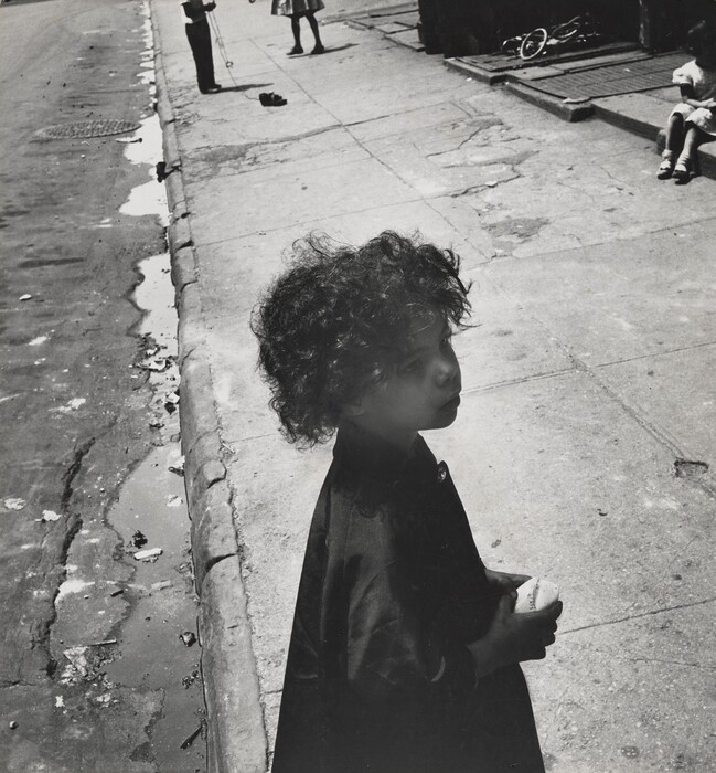 This is a photo of a city street scene. The image shows an urban sidewalk with a young child holding a ball. The child has curly hair and is deep in thought. The sidewalk is cracked, indicating wear. Toys or bicycles are visible on the side. In the background, two other children are seen; one skips a rope and the other sits further back. The angle of the photo emphasizes the child's expression and the texture of the surroundings. The photo is in black and white.