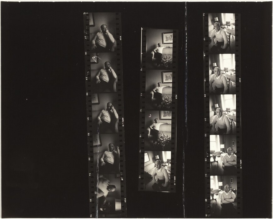 The contact sheet displays a series of photographs of a man seated in different poses. He is shown from the waist up to full body in some images. The man has a bald head and is wearing a light-colored sweater and dark pants. He appears relaxed and contemplative. The background includes framed artwork on the walls, a coffee table with various objects, and a room with large windows letting in natural light. Additional elements like a sofa and a lamp are visible in a few frames.