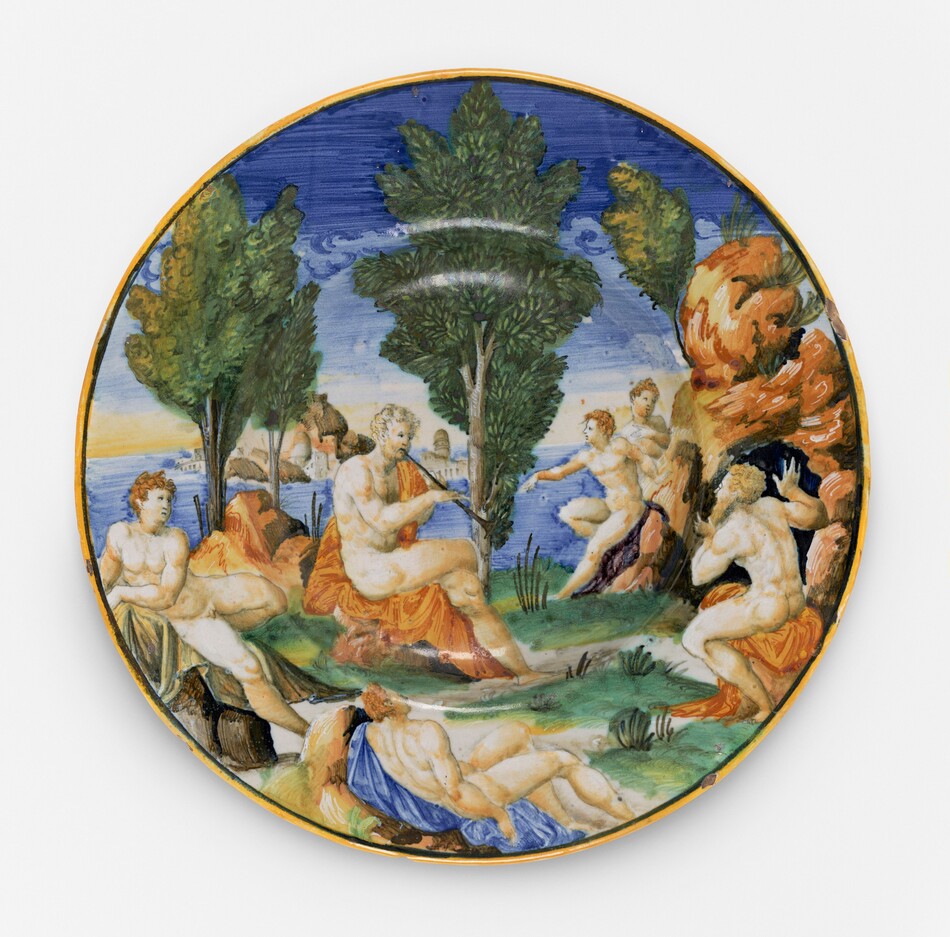 This is a painting of a pastoral scene with figures around a central tree on a circular ceramic dish. The image depicts several nude and partially draped classical figures in a lush landscape. At the center, a figure is playing a lute, surrounded by others who appear to be listening or engaging in their own activities. The background includes greenery, distant architectural structures, a body of water, and a sky with soft clouds. This is in the style of Renaissance art.