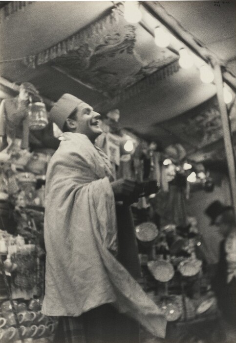 The image shows a man in a three-quarters profile, displaying part of his torso. He stands with a slightly tilted head and a wide smile on his face. The man wears a traditional fez and is draped in a white garment over his shoulders. He is holding a black object. The surroundings suggest a lively environment, possibly a market or stall, with shelves lined with various goods and items hanging around. The background is decorated with patterned fabrics or tapestries on the ceiling, illuminated by light bulbs.