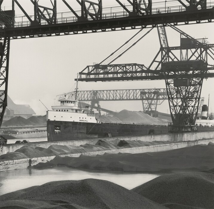 The image displays an industrial scene featuring a large cargo ship named "MARHILL" docked at a port. The view is from slightly above water level, showing the ship, industrial cranes, conveyer systems, and mounds of material. The style is detailed, depicting the geometric lines of the cranes and the ship's hull. The color scheme is monochromatic with shades of gray and black. The background includes more cranes and industrial structures, and the sky is overcast, providing diffused lighting.