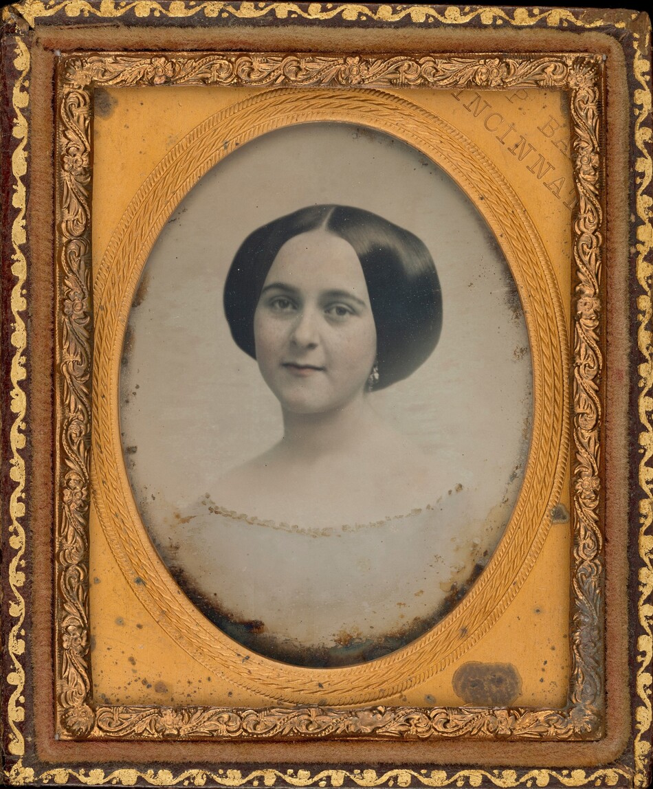 The image shows a portrait of a woman, capturing her from the shoulders up. She is positioned directly facing the camera with a slight tilt of her head. The woman has smooth, dark hair styled in a middle part, with her hair sleekly wrapped over her ears. Her facial features include dark eyes and full lips. She is wearing an off-shoulder garment, possibly a dress or blouse, embellished with a delicate trim. The woman is also adorned with earrings. The photograph is enclosed in an ornate gold-tone frame with intricate floral and scrollwork details on the outer edge. The background within the frame appears neutral and unadorned.