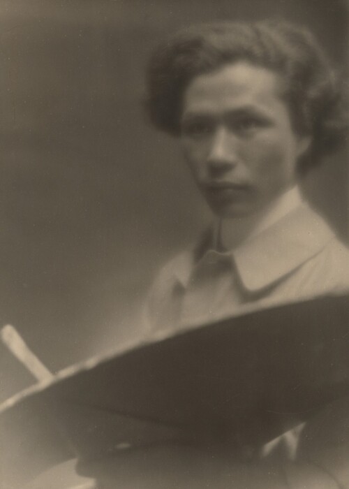 The image shows a close-up of an individual's face and part of the upper body. The person is in a seated position, facing slightly to the side with their face turned towards the camera, giving a partial profile view. The person has a full head of dark hair styled in a wavy manner and is wearing a light-colored collared shirt. In front of them is a large, round object resembling an artist's palette. The background is a plain, smooth surface with a neutral tone.