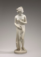 A nearly nude woman is carved from cream-white marble in this freestanding sculpture. In this photograph, her body faces us, and she looks to our right in profile. She has a sloping forehead and straight nose, full, closed lips, and a rounded chin. Her curly hair is pulled back but ringlets fall down the back of her neck, and tendrils curl alongside her ears. She holds her left arm, to our right, across her torso so that hand brushes her right breast. She holds drapery to her groin with the other hand, and the cloth puddles behind her feet. She steps slightly forward onto her right leg but her knees are together so the toes of her left foot brush the round, disk-like base on which she stands. The background fades from nickel gray across the top to fog gray along the bottom.