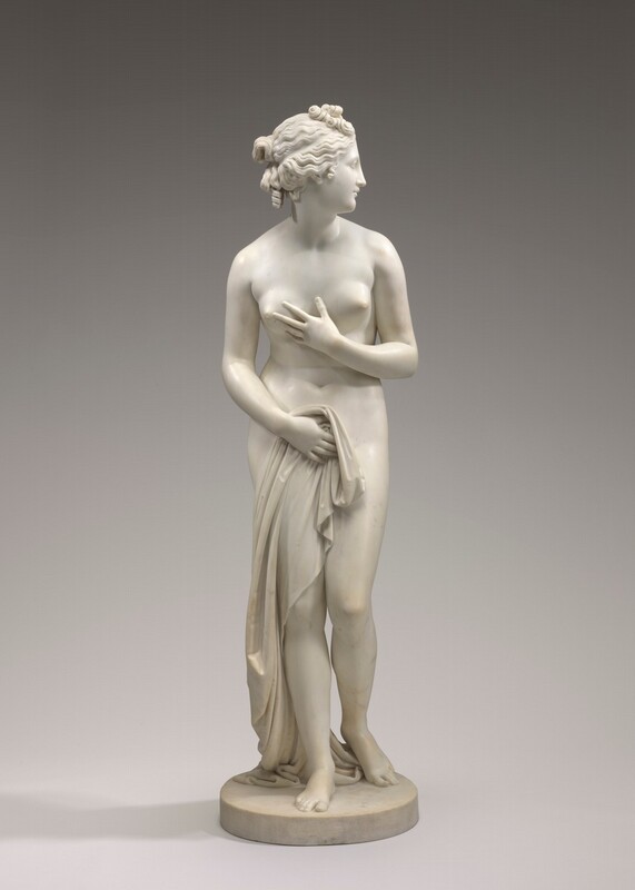 A nearly nude woman is carved from cream-white marble in this freestanding sculpture. In this photograph, her body faces us, and she looks to our right in profile. She has a sloping forehead and straight nose, full, closed lips, and a rounded chin. Her curly hair is pulled back but ringlets fall down the back of her neck, and tendrils curl alongside her ears. She holds her left arm, to our right, across her torso so that hand brushes her right breast. She holds drapery to her groin with the other hand, and the cloth puddles behind her feet. She steps slightly forward onto her right leg but her knees are together so the toes of her left foot brush the round, disk-like base on which she stands. The background fades from nickel gray across the top to fog gray along the bottom.