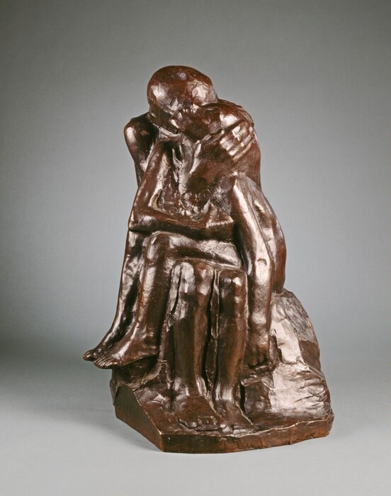 The sculpture depicts two human figures engaged in an embrace. The figures are rendered realistically with detailed anatomical features. The sculpture is a rich, brownish color, possibly made of bronze or a similar metal. The surface is smooth yet textured, with polished areas and subtle details. The composition emphasizes the connection and closeness between the figures, highlighting their physical proximity and complementary forms.