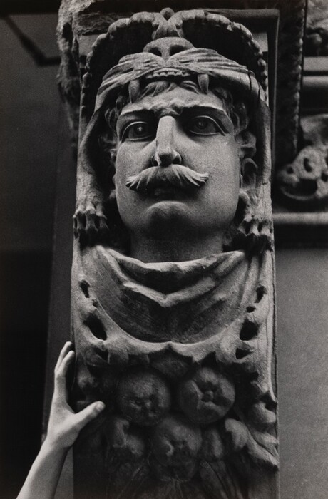 The sculpture features the face of a man with a mustache, strong facial features, and curly hair framing his face. The upper part of the sculpture includes a headdress or helmet with a stylized animal motif, possibly a lion's head with an open mouth. The face is serious and frontal, and beneath the chin, there are decorative elements resembling clusters of fruits or rounded objects. The material seems to be stone, apparent from its rough, weathered texture and monochromatic hue in the black and white photograph. A human hand is visible at the lower left for scale, indicating the sculpture's size. The stone surface shows a mix of smooth and rough areas, highlighting the sculpture's intricate details.
