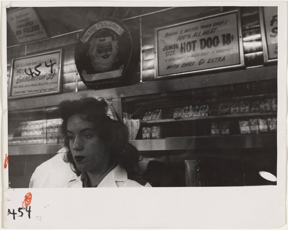 This is a photograph of the interior of a diner or food establishment. The image captures a section of the diner with behind-the-counter shelves stocked with food products. Above, there are multiple signs displayed, including advertisements for jumbo hot dogs and sandwiches. The space has a festive touch due to the "Merry Christmas" sign with a Santa illustration visible. The black-and-white nature of the photograph gives it a vintage feel, highlighting the textures of the metal surfaces and the visual contrast between the light and dark areas.