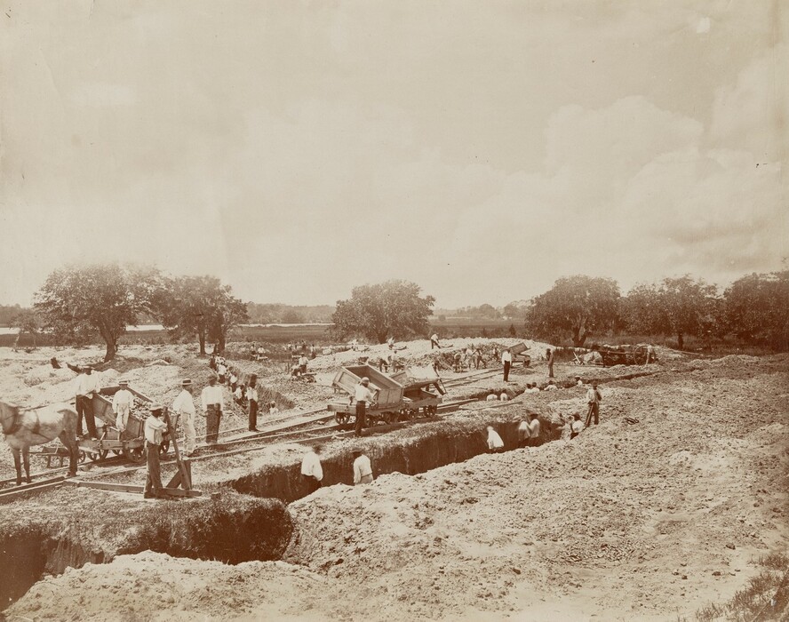 The image shows a construction area with workers digging and handling materials. In the background, there are trees indicating a wooded area. Some figures are standing or moving near tracks, with a horse-drawn cart transporting goods. The image is in sepia tones, giving it an antique quality.