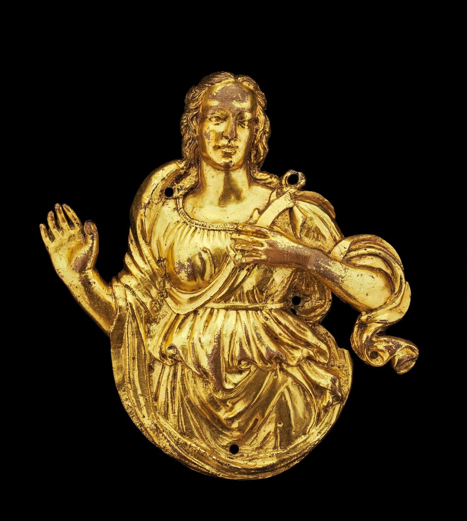 The sculpture depicts a figure in a classical pose. The subject is a person clad in draped clothing, with the right hand raised and the left hand positioned closer to the chest. The sculpture is made from a golden material and has a smooth surface with detailed folds in the clothing. The figure appears to be a bust with an intricate robe. The material gives the sculpture a polished and luminescent quality. It does not have a visible base. The features of the figure, such as facial expressions, hands, and hair, are skillfully rendered.