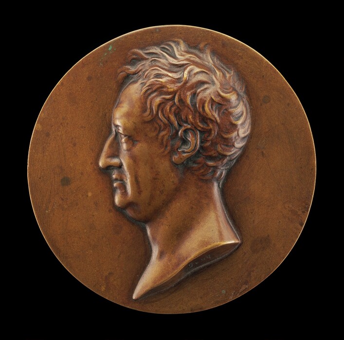 The sculpture is a round medallion depicting a side profile of a man's head and upper neck in relief. The subject has a prominent nose, firm chin, and curly hair. The sculpture is made of a smooth, reddish-brown material, likely bronze. There are small spots on the surface. The background is a simple, flat surface. The overall effect is a classic profile portrait similar to ancient coins or medals.