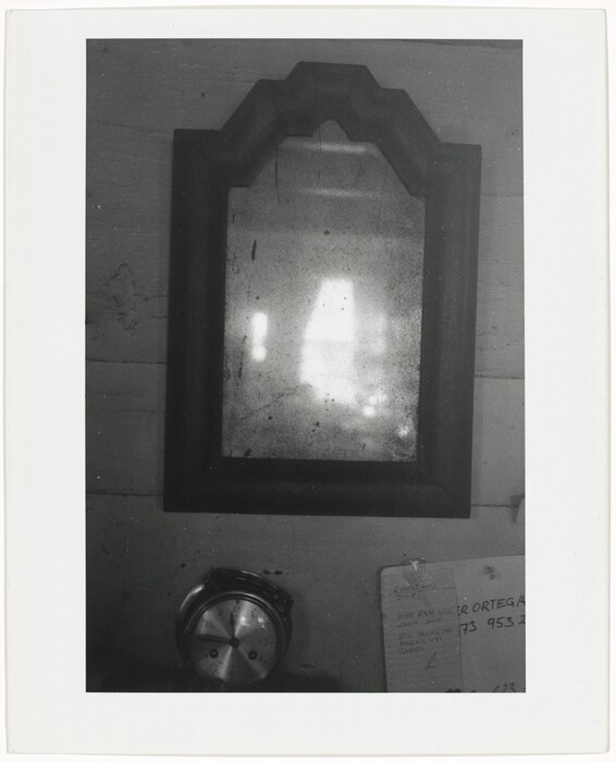 The image shows a large, ornately framed mirror with a vintage appearance at the center. The overall color palette is grayscale, typical of a black-and-white photograph. The mirror reflects light and shadows, and below it to the left is a circular metal object. To the right of the metal object is a piece of paper with text in the lower right corner. The background is a wooden wall.