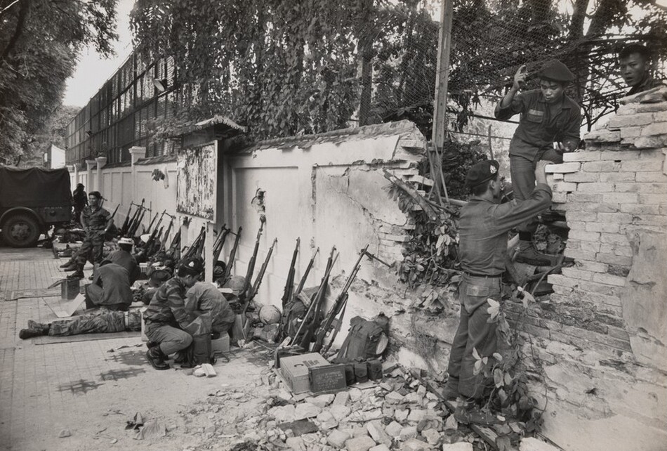 This grayscale photograph shows a group of soldiers on a sidewalk next to a crumbling brick wall. Several soldiers are seated or crouched on the ground with rifles propped up against the wall. One soldier is standing and assisting another soldier climbing over a damaged section of the wall on the right. In the background on the left, a large vehicle is parked under a canopy of trees. Debris and military gear are scattered in the area. Vegetation is visible along the top edge of the wall, and fallen bricks are scattered across the pavement.