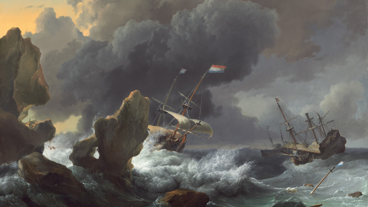 Beyond several craggy boulders that loom in the lower left corner of this horizontal painting, three sailing ships pitch wildly in crashing waves beneath towering clouds. At the center, a large ship tips sharply to our right with billowing ivory sails and two red, white, and blue striped flags whipping in the wind. White spray kicks up against the side of the boat and in the waves surrounding it. The sails of the second ship, to our right, are furled except for one that crashes down onto the deck. Tiny people scurry around inside the ship, which tilts steeply up on a high wave. The third ship floats beyond this, its sails also tied up. The top of a tall wooden mast along with a broken wooden pole poke up from emerald-green waves in the lower right corner, near a barrel and two bundles wrapped in cloth and tied with rope that bob nearby. One of the brown, jagged rocks to our left nearly spans the height of the painting while others jut from the water like crooked teeth. A bank of billowing, slate-gray clouds at the center of the sky separates a fog-gray sky and puffy clouds to our right from a patch of golden sunlight to our left, in the upper corner of the canvas. The artist signed and dated the work as if written on a rock at the bottom center of the canvas, “LBackh 1667.”