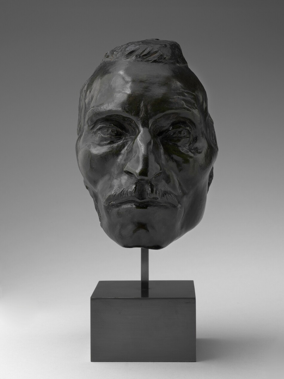The sculpture is a bust depicting the head and face of a man. The figure has an elongated face with prominent cheekbones, a straight nose, and a notable mustache. The hair is short and combed back. The sculpture appears to be cast in a dark-colored metal, possibly bronze, giving it a shiny, reflective quality. The head is mounted on a simple, square base with a small vertical rod supporting the face, creating an impression of the head slightly elevated above the base. The expression on the face is neutral.