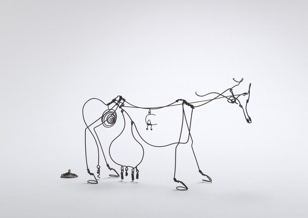 The sculpture is made of thin black wire and depicts the abstract outline of a cow. The wire is skillfully twisted and looped to outline the cow's anatomy, including its head, with ears and a nose, two long horns, an udder that hangs down towards the ground, and curled wire for its legs. The head of the cow faces the right. Behind the cow, on the ground to the left, is a small pile of twisted wire that resembles a pile of feces.