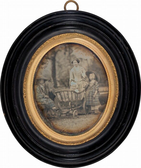 The image features an oval-shaped photograph encased in a black and gold frame. In the photo, a group of people is in an outdoor setting. In the center, a woman stands next to a child. In front of them is a wooden pushcart with a baby seated inside. Two other people are partially visible to the side. The background is blurred, suggesting a natural setting with trees or foliage. The color scheme is sepia-toned, indicating an aged photograph.