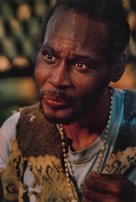 The image depicts a man's face and upper body, with a focus on his left side. He is seated and turned slightly to his right. The man has a beard, dark curly hair, and a contemplative expression. He is wearing a light blue shirt under a brownish patterned vest or jacket. Around his neck, he wears a string of white beads. The background is blurred with a mix of light and shadow, suggesting an outdoor setting with foliage or trees in the distance.