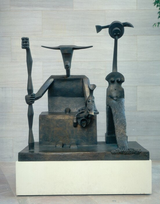 Two abstracted creatures, one resembling a goat holding a staff and the other a stylized mermaid, sit facing us on on a rectangular base in this free-standing bronze sculpture. The head of the goat creature, to our left in this photograph, has wide horns to either side of a ridge suggesting a heavy brow. The triangular head tapers down to a ring that suggests the muzzle of the goat. The body is made up of broad geometric shapes. There is a stylized phallus where the body meets the block we read as a lap, and two rounded forms suggestive of human feet protrude from the base of that block. The creature reaches out with his right arm, to our left, to wrap a large, human fist around the staff. The staff swells and narrows in angular hourglass shapes and is topped by what could be a rectangular, abstracted face with two eye holes, a flattened nose, and a slit for a mouth. The goat’s other hand rests near that knee, and in it he holds a creature about the height of the goat’s torso. That smaller creature has a scooped, cup-like form where the head would be, a long neck, and two mounds reminiscent of breasts above a textured fish’s tail. Below, as if affixed to the front of the goat’s lap, a disk-like face has two rings for eyes and a tongue protruding from a round mouth. Other forms over the forehead and one next to the face read as flattened hands or claws. Next to the large goat creature, to our right, the second, tall creature has a head made up of a disk with two mounds for eyes and a beak stacked in front of a second, larger disk. A form like an abstracted fish or arrow moves through or behind her head. The creature has a long, slender neck and an armless torso, shaped roughly like the body of a violin. The torso has rounded shoulders, two mounds to suggest breasts, and a hole for a belly button. Her textured fish’s tail curves down so the fin falls slightly over the front of the base on which they sit. The bronze surface of the sculpture varies from dark brown to faint gold where the light catches it. The sculpture and bronze base sit on a white platform. The floor beneath is pale pink, shiny marble and the wall behind is made up of stone panels streaked with gray.