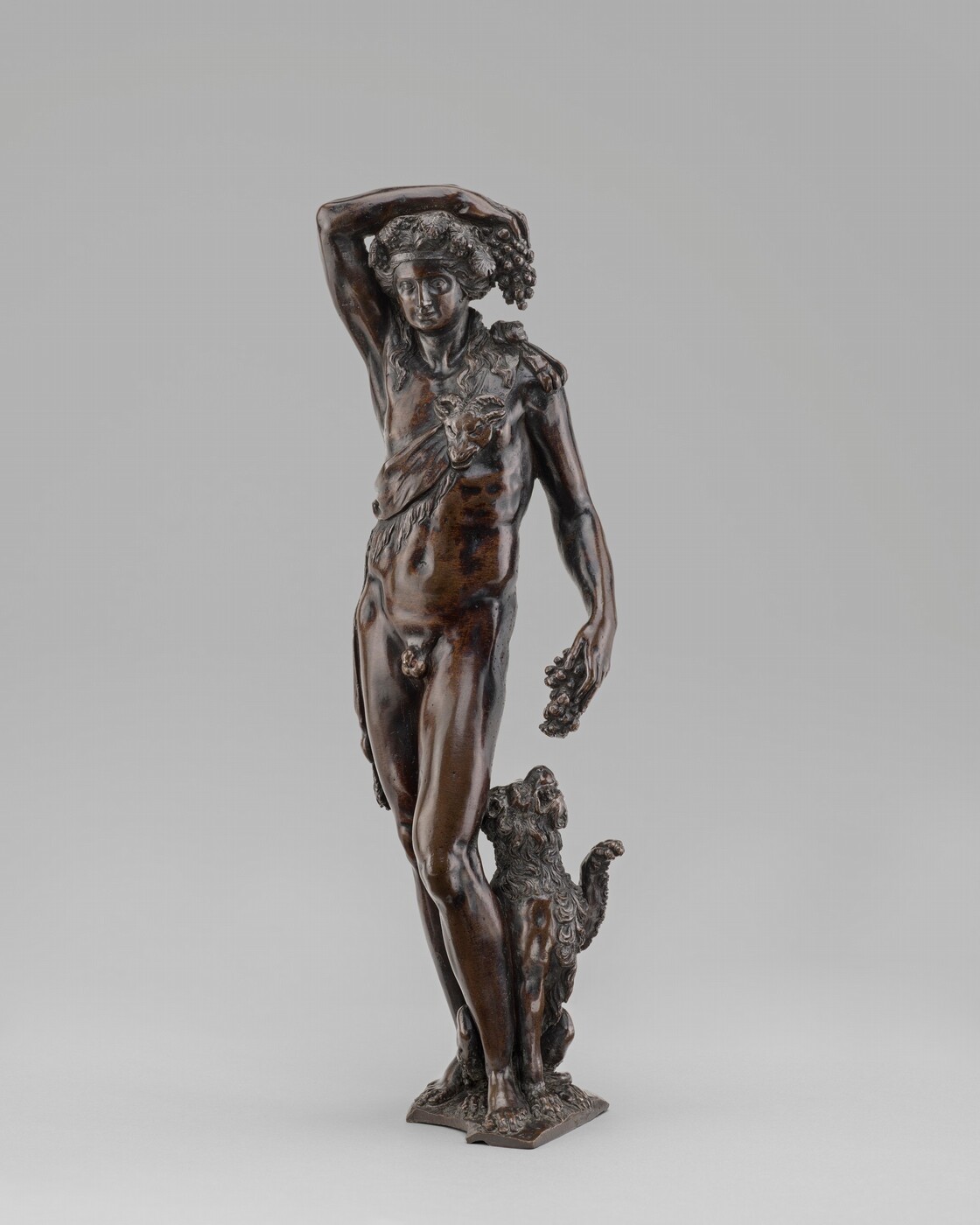 The sculpture features a male figure with one arm on his head and the other holding grapes, accompanied by a small animal. The figure wears a garment with a goat's head motif and the sculpture is made of a dark brown material, likely bronze. The animal's fur is detailed, and the sculpture stands on a textured base.