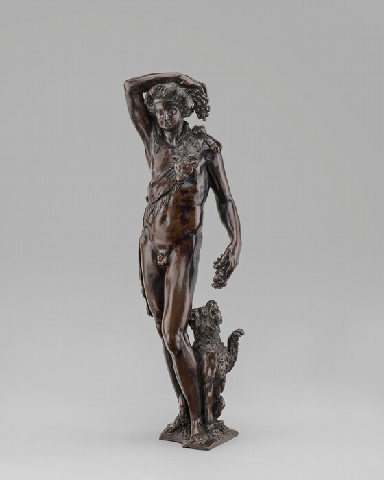 The sculpture features a male figure with one arm on his head and the other holding grapes, accompanied by a small animal. The figure wears a garment with a goat's head motif and the sculpture is made of a dark brown material, likely bronze. The animal's fur is detailed, and the sculpture stands on a textured base.