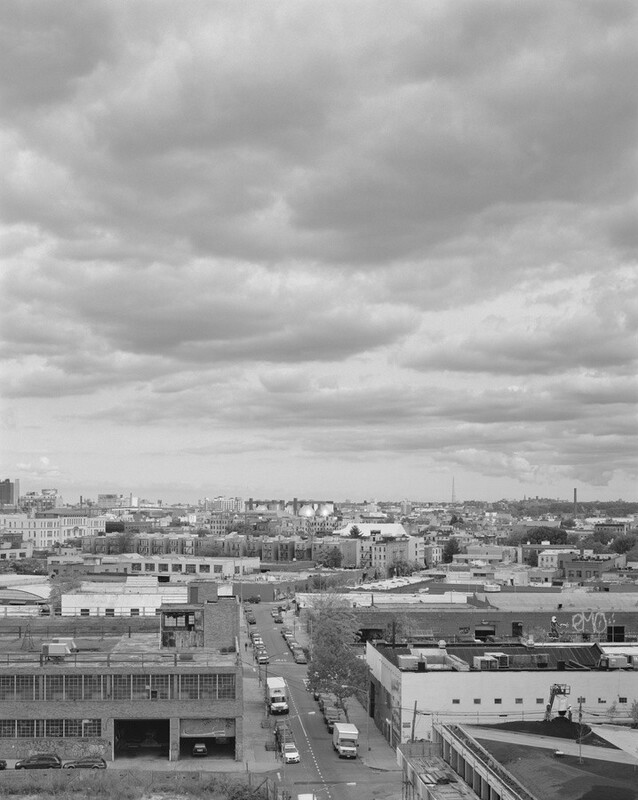 This grayscale photograph shows an elevated view of an urban landscape. The horizon is in the middle of the image, dividing the cloudy sky and the cityscape. The sky contains thick, textured clouds. The city below has industrial and residential buildings extending into the distance. A road runs straight through the landscape, with parked vehicles on both sides.